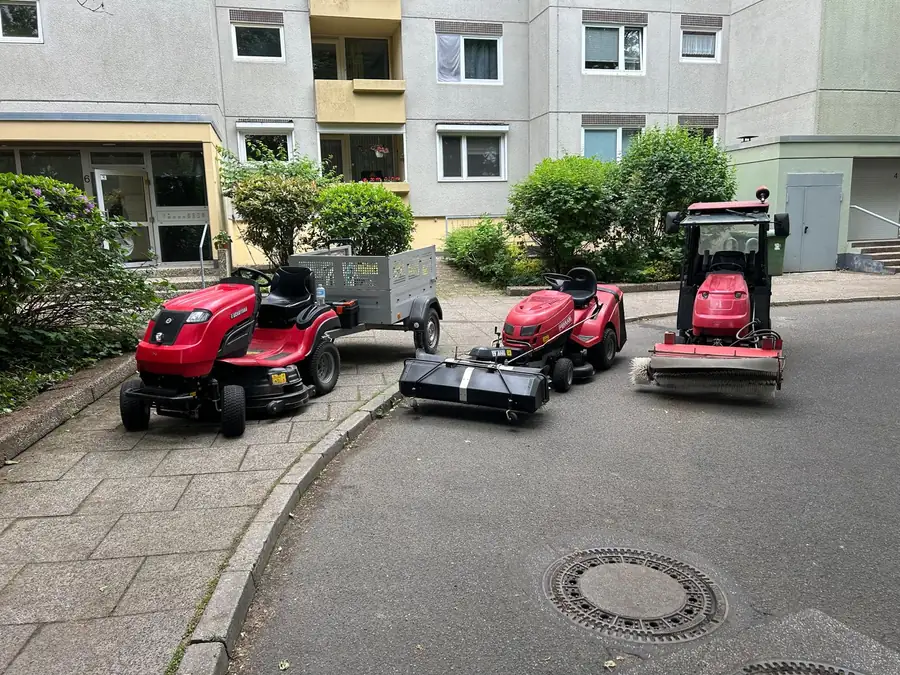 Drei rote Aufsitzmäher und eine Kehrwalze im Hof vor einem hellen Wohnblock