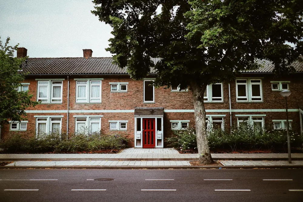 Zweigeschossige Klinker-Wohnanlage mit roter Haustür, Hecke und großem Baum an einer ruhigen Straße